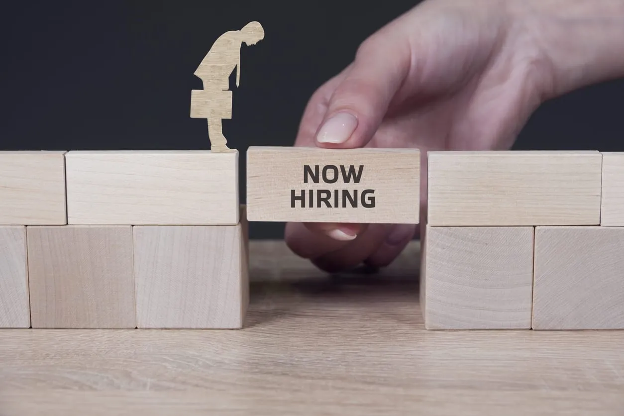 Une main place un bloc de bois portant l’inscription « Now Hiring » entre deux espaces, créant ainsi un passage pour une figurine munie d’une mallette – une métaphore de l’headhunting inversé.